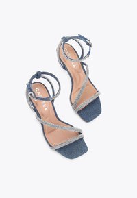 Sandały na obcasie z denimu mają kwadratowy czubek, krzyżujące się paski z akcentami z cyrkonii oraz lekko wyściełaną beżową wkładkę.