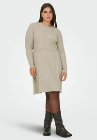 Vestido de punto de color gris claro, con mangas largas, escote redondo y falda acampanada. Llevado con botas negras hasta el tobillo y medias oscuras.