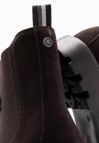 Botas de tobillo de ante marrón con un panel de punto texturizado, suela de goma negra y talón reforzado. Presentan una tira de agarre a rayas y un detalle del logo.