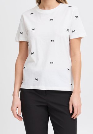 Camiseta blanca de manga corta con pequeños lazos negros, usada por una persona con las manos relajadas a los lados, combinada con pantalones negros.