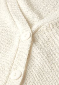 Cardigan en tricot blanc avec un col en V, un tissu texturé et deux grands boutons ronds blancs sur le plastron avant.