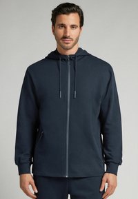 Sudadera de color marino hecha de tejido suave con cremallera frontal, capucha con cordón y bolsillos laterales. Presenta un corte holgado y puños acanalados.