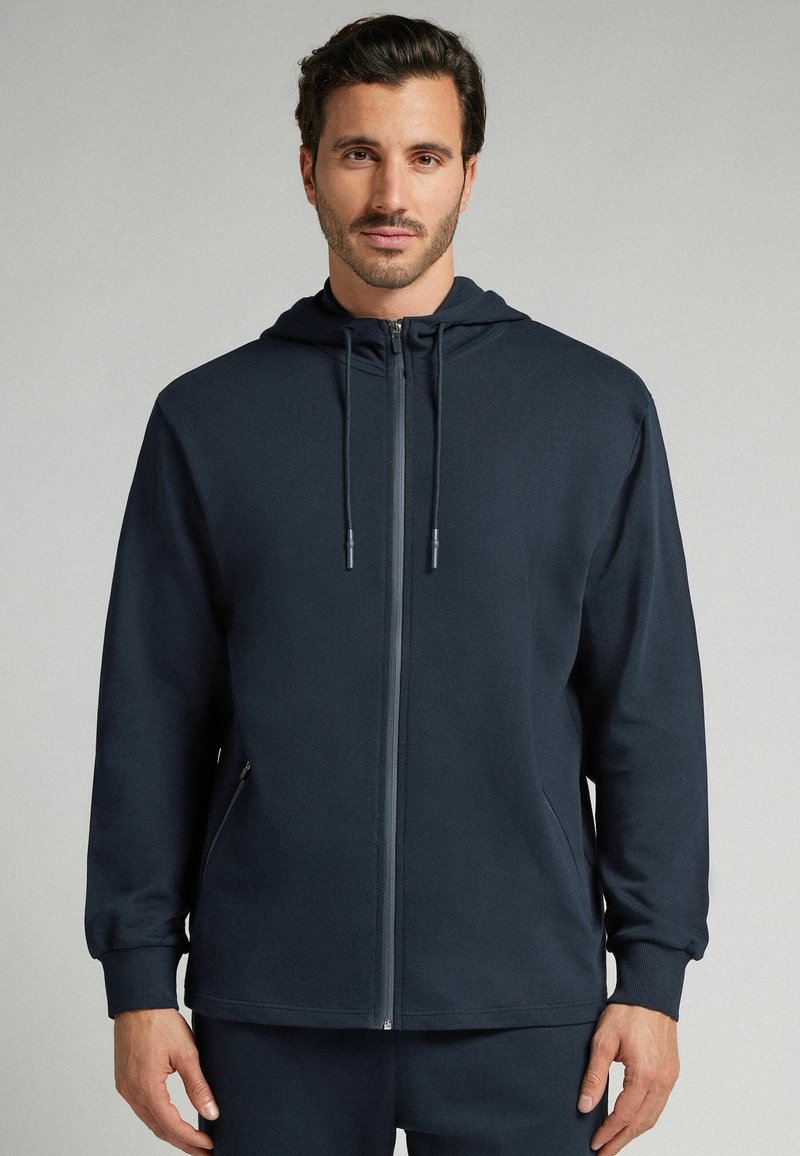 Sudadera de color marino hecha de tejido suave con cremallera frontal, capucha con cordón y bolsillos laterales. Presenta un corte holgado y puños acanalados.