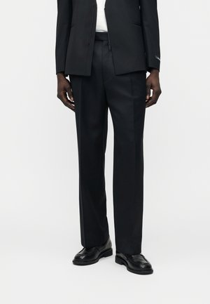 Meesmodell, kes kannab musta kohandatud pükse, musti nahkjalatseid ja musta jopet valge pluusi kohal, seisab ühevärvilise tausta ees.