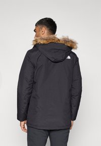 Zwarte parka jas met nep bonttrand op de capuchon, met een logo op de achterkant, praktische ontwerp en een gladde textuur.