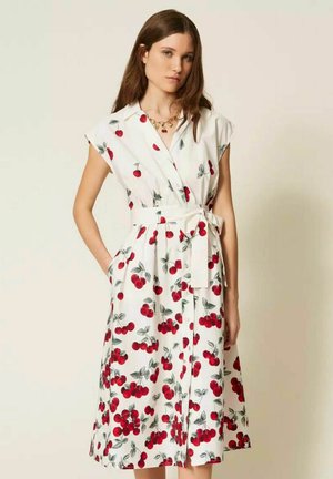Giovane donna che indossa un abito bianco senza maniche con stampa di ciliegie rosse e cintura annodata in vita, in piedi con una mano in tasca.