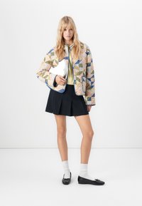 Giubbotto bomber trapuntato con motivi geometrici multicolori, gonna nera plissettata, camicia bianca e ballerine nere con fiocchi. Clutch bianca.