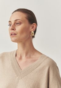 Beige V-nyakú pulóver, texturált varrási részletekkel, arany háromszög fülbevalókkal viselve, semleges háttér előtt.