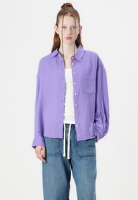 Light purple button-up shirt made of textured cotton, featuring a chest pocket and relaxed fit, paired with loose blue denim pants.