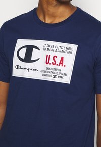 Muž v námořnickém modrém tričku Champion s bílým nápisem: "Je třeba trochu víc, abyste se stali šampionem. U.S.A. autentické sportovní oblečení."