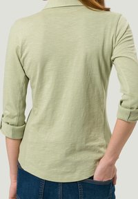 Top verde chiaro a maniche lunghe con colletto e maniche arrotolate, caratterizzato da una texture liscia e una vestibilità leggermente comoda.