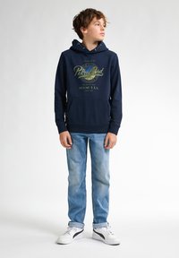 Sudadera azul marino con estampado gráfico, bolsillo tipo canguro en la parte frontal y capucha con cordón, combinada con jeans azul claro y zapatillas blancas.
