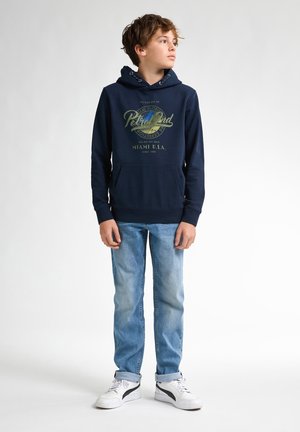 Felpa blu navy con stampa grafica, tasca a canguro frontale e cappuccio con cordino, abbinata a jeans azzurri e sneakers bianche.