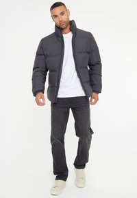 Sort puffjakke med høj krave, quiltet design og elastiske manchetter, parret med en hvid t-shirt og mørke jeans. Lyse sko.