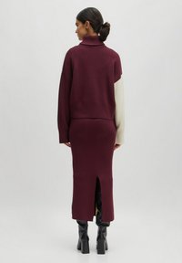 Burgunderfarbenes Strickkleid mit hohem Kragen, gerippten Details und einem seitlichen Schlitz. Verfügt über kontrastierende cremefarbene Ärmel und kniehohe schwarze Stiefel.