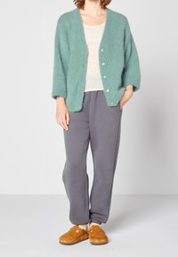Un cardigan moelleux vert menthe avec un col en V et de gros boutons, associé à un pantalon de survêtement gris et des sabots en daim marron. Tenue simple et confortable.