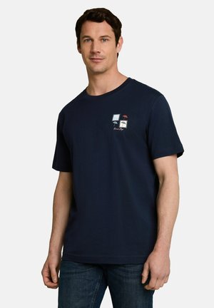 Man draagt een donkerblauw t-shirt met een kleine paraplu en een vierkante afbeelding op de borst, gecombineerd met blauwe jeans, poseren voor een effen witte achtergrond.