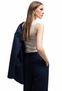 Marineblaue Streifenanzugjacke über einer Schulter drapiert, kombiniert mit einem beigen gerippten Tanktop. Langes Haar und goldene Creolenohrringe sichtbar.
