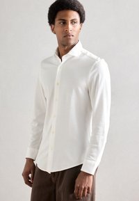 Valge pikkade varrukatega särk kraega kaelusega, millel on nööbitav eesmine osa, pehme kangas ja kergelt kaardus serv; sobitatud pruuni pükstega.