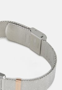 Bracelet de montre en mailles argentées avec un fermoir métallique. Présente un design texturé et un petit accent doré près du fermoir. Matériau en acier inoxydable.