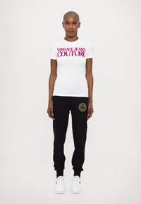 Hvit T-skjorte med fet rosa "VERSACE JEANS COUTURE" tekst, kombinert med svarte joggebukser med et gull logo på venstre lår og hvite sneakers.