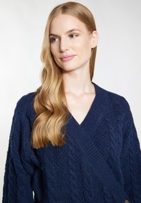 Marinblå stickad cardigan med v-ringning, strukturerad kabelmönster och breda ribbade ärmslut. Har en enkel knappstängning framtill.