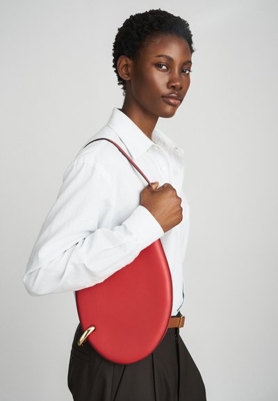 Bolsa oval vermelha com uma textura de couro suave e um detalhe em tom de ouro. Usada com uma camisa de riscas claras e um cinto castanho.