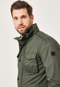 Green cotton jacket features a high collar, multiple front pockets, and a textured design with contrasting stitching and a logo patch.