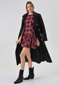 Donna che indossa un lungo cappotto nero sopra un vestito a quadri rosso e nero con orlo arricciato e stivali neri alla caviglia, in piedi contro uno sfondo bianco.