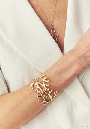 Goldenes armband in Korallenform am Handgelenk und passende goldene Korallen-Anhänger-Halskette, getragen über einem weißen Blazer.