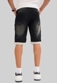 Shorts en denim noir avec des bords roulés, présentant un délavage teinté, deux poches arrière et des détails de couture visibles. Portés avec des baskets blanches.