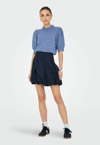 Modrý pletený sveter s nafúknutými rukávmi, v kombinácii s námornícky modrou, skladanou sukňou. Modelka má na sebe tmavé topánky a biele ponožky po členky.