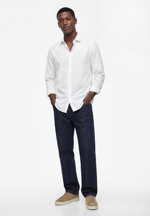 Mann steht mit den Händen in den Taschen, trägt ein weißes Hemd mit Knöpfen, dunkelblaue Jeans und beige Sneaker vor einem einfarbigen weißen Hintergrund.