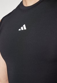 Black athletic sleeveless top made of smooth, stretchy fabric. Features a white logo with three stripes on the upper left chest.