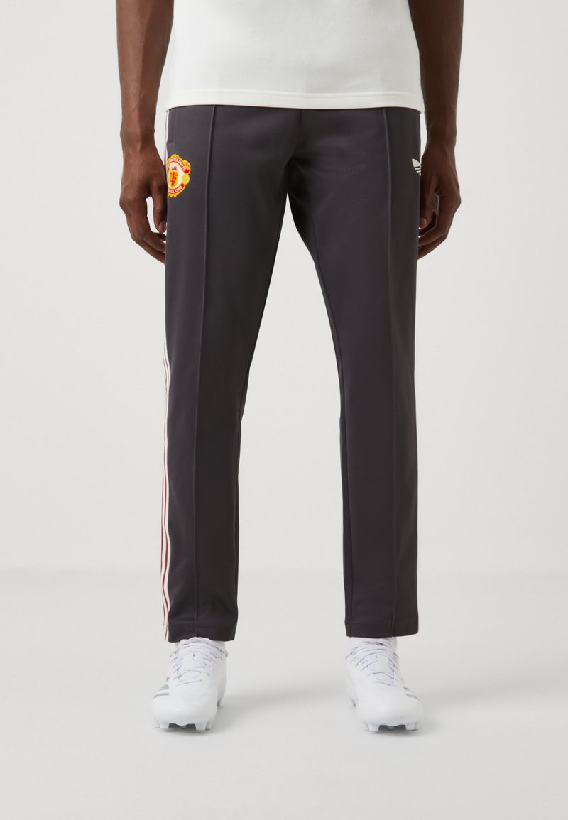 Pantalon de sport gris foncé en tissu lisse, avec des rayures blanches et rouges sur les côtés et un logo brodé sur la cuisse supérieure.