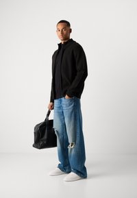Homme portant un sweat zippé noir, un jean bleu déchiré ample, des baskets blanches, tenant un sac en cuir noir, se tenant contre un fond clair uni.