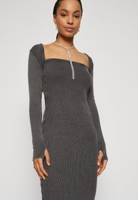 Robe ajustée à manches longues en tissu côtelé gris foncé. Présente un décolleté carré et des poignets avec trous pour les pouces. Détails remarquables comprenant une pierre précieuse.