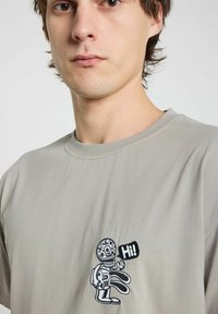 T-shirt en coton gris avec un design brodé noir représentant un squelette et un lapin, contenant le mot "Salut !" dans une bulle de dialogue.