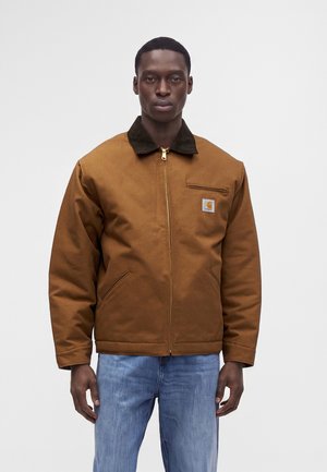Homme portant une veste marron à fermeture éclair avec un col foncé et le logo Carhartt, associée à un jean bleu clair, debout devant un fond uni.