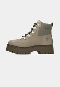 Beige semsket ankelstøvel med et teksturert grått panel, metalløyer og en robust brun plattformsåle med dypt mønster.