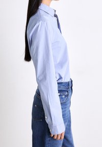 Camisa de botones color azul claro hecha de algodón, con mangas largas, un cuello clásico y una textura suave, combinada con jeans de mezclilla azules.
