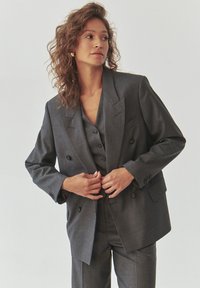 Grijze dubbelknoopsblazer met notch-revers, knoopdetails en een bijpassend vest eronder. Gladde textuur, getailleerde pasvorm.