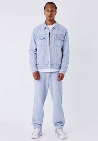 Lichtblauwe denim jas en broek set met een subtiele patroonstructuur, voorzakken en een aansluitend ontwerp. Gedragen over een wit shirt.