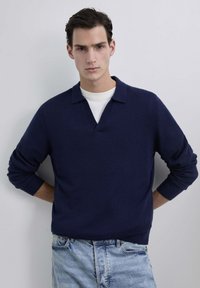 Suéter azul marino con cuello en V y cuello, hecho de material suave, con mangas largas y un ajuste relajado. Llevado sobre una camisa blanca.