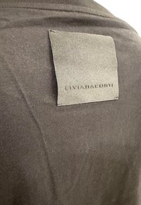 Sort t-shirt med korte ærmer, der har et rektangulært stofmærke på bagsiden med "LIVIANACOM" præget. Glat tekstur, ingen mønstre.