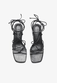 Schwarze Ledersandalen mit quadratischer Zehenpartie, die mit überkreuzten Schnürungen und dünnen Riemen versehen sind. Glatte Textur mit einem minimalistischen Design.