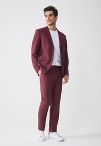 Burgundy suit with a single-button blazer and matching trousers, paired with a white t-shirt and white sneakers. Smooth fabric texture.