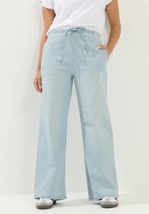 Person iført lyseblå bukser med vide ben og snøre i taljen, hvid T-shirt og hvide sneakers, stående foran en ensfarvet baggrund.