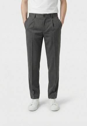 Mann iført mørkegrå plisserte bukser med lommer, hvite sneakers og en hvit t-skjorte, stående med hendene i lommene.