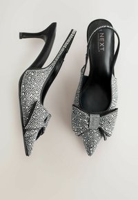 Mustade slingback kõrged kontsad, millel on terav nina, hõbedaste rhinestone'idega kaetud kangas ja ees kaunistav lips. Kunstnahast läikiv äär.